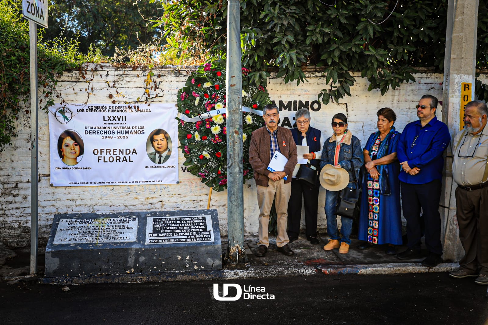 Personas de pie en una banqueta junto a una ofrenda florar con una lona