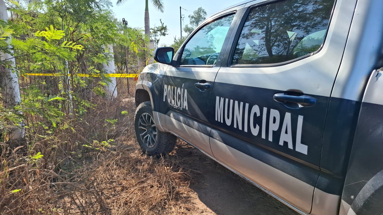 Unidad policiaca en una calle de terracería y al fondo se observa una cinta amarilla de precaución