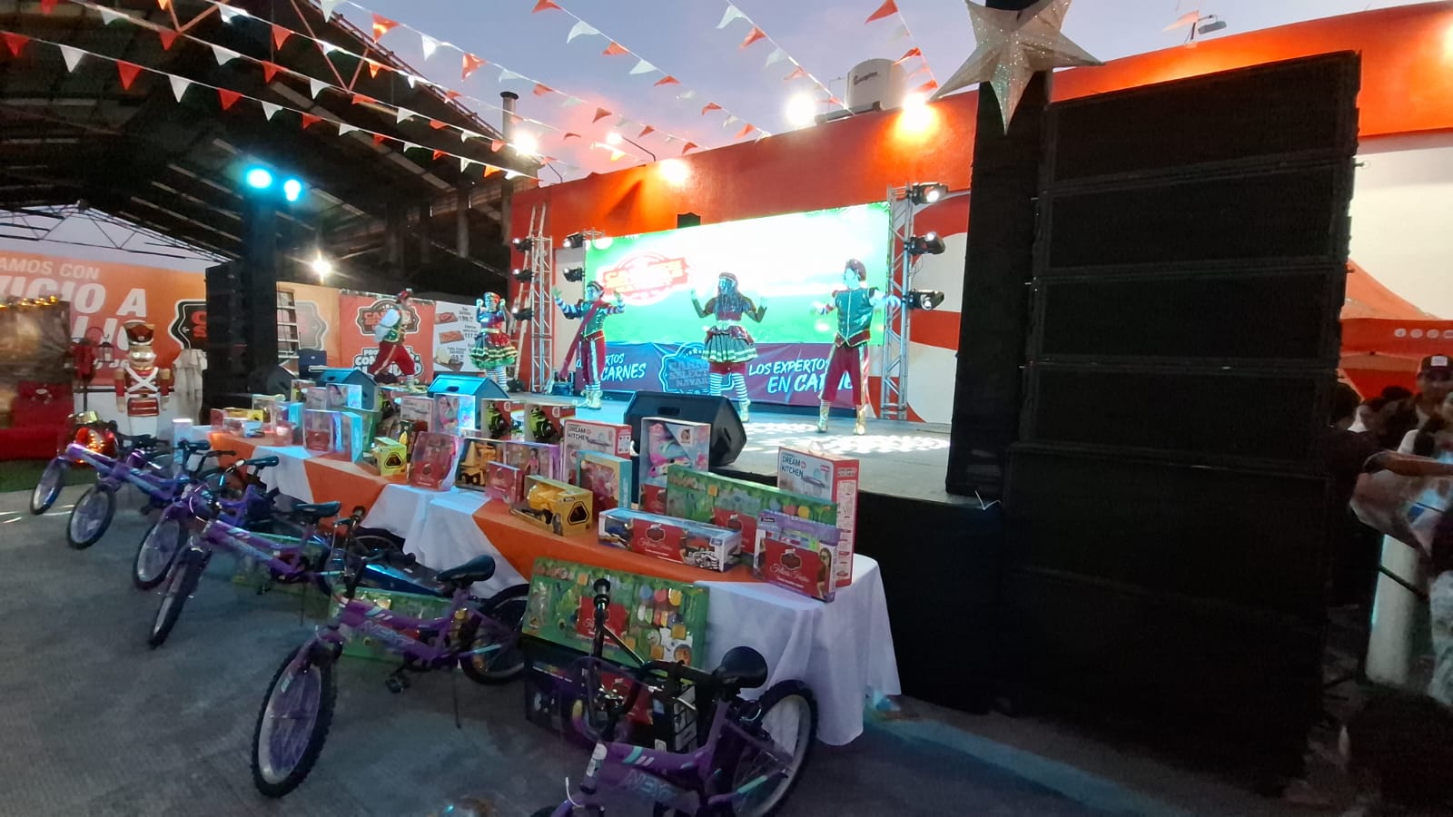 Escenario con decoración navideña y bailarines arriba de el, debajo una mesa con variedad de juguetes y bicicletas