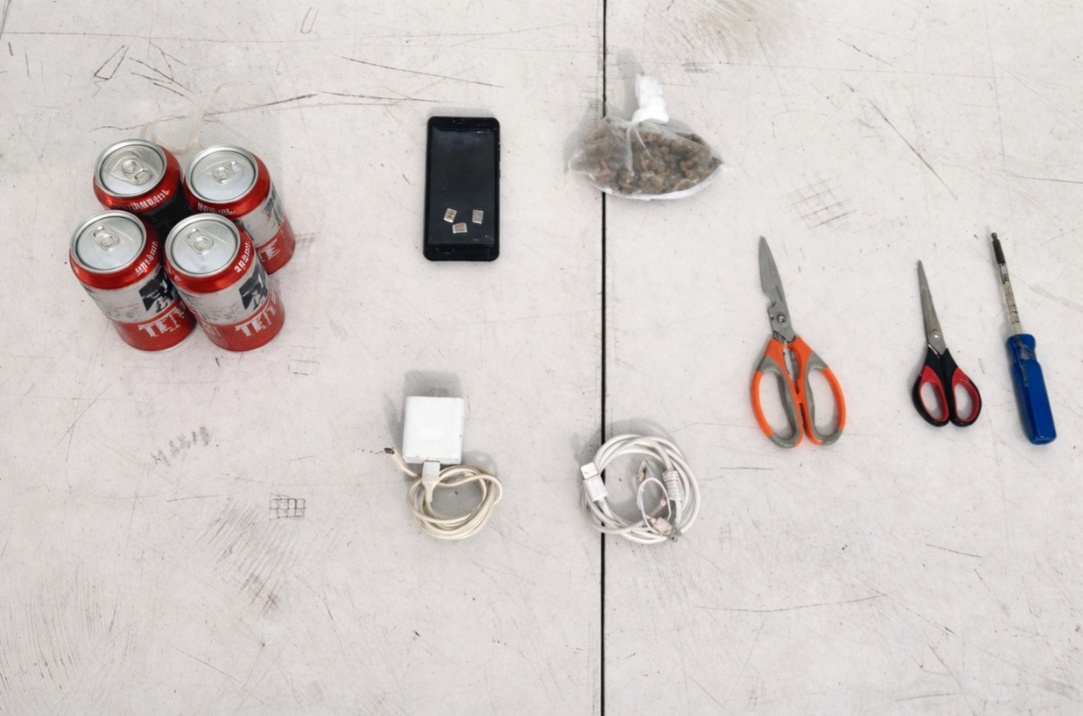 Cuatro latas de cerveza, tijeras, cargadores u un celular sobre una mesa blanca