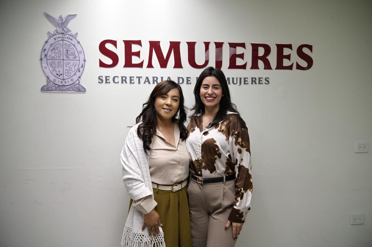 Dos mujeres abrazadas viendo hacia el frente sonriendo y atrás un letrero que dice SEMUJERES