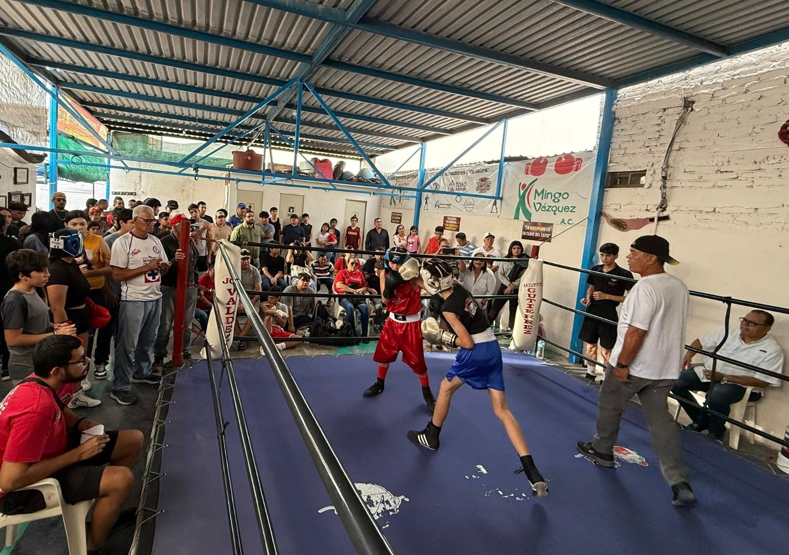 Boxeadores en un ring peleando, con un réferi observando el combate, debajo del ring publico observando y animando a los boxeadores