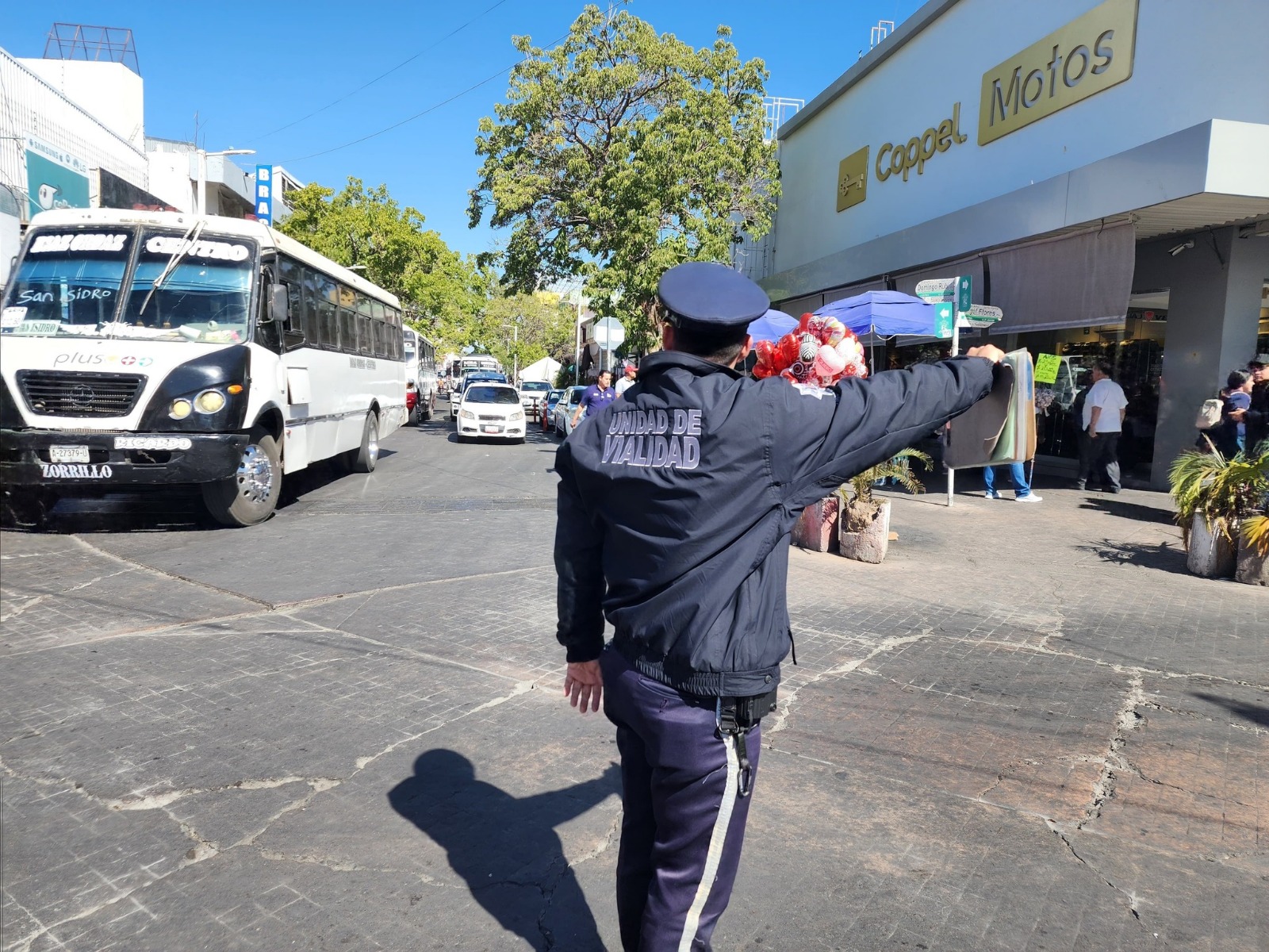 Elemento policiaco en medio de la calle, se observa un camión y carros y establecimientos