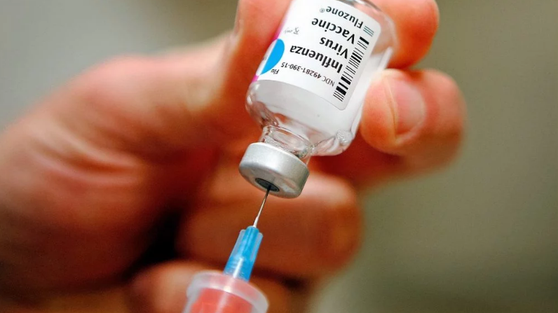 Manos sosteniendo una aguja de una jeringa que está introducida en un contenedor de medicamento inyectable con una etiqueta que dice influenza
