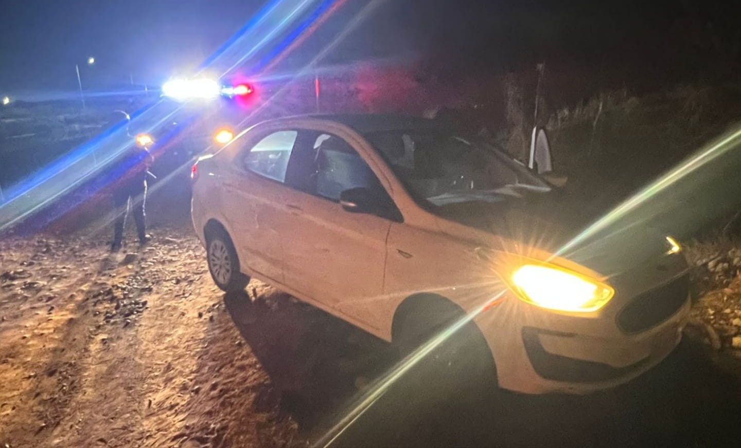 Vehículo blanco con las luces prendidas y al fondo unidad policiaca con las luces encendidas