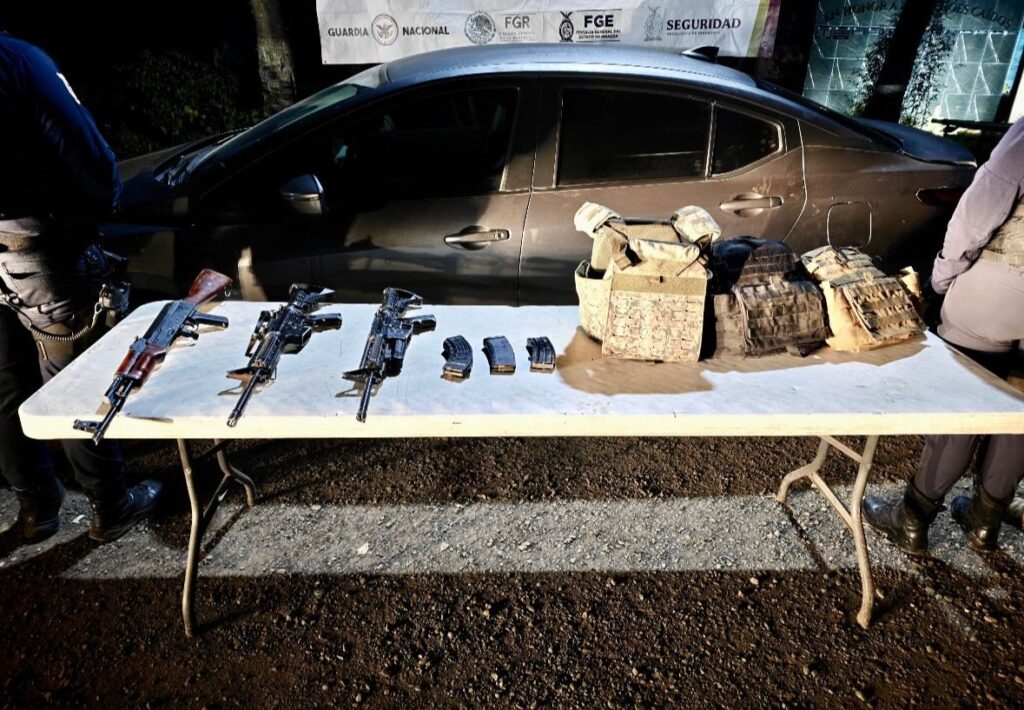 Mesa con armas, cargadores y más equipo que fue asegurado, al fondo un vehículo y una lona