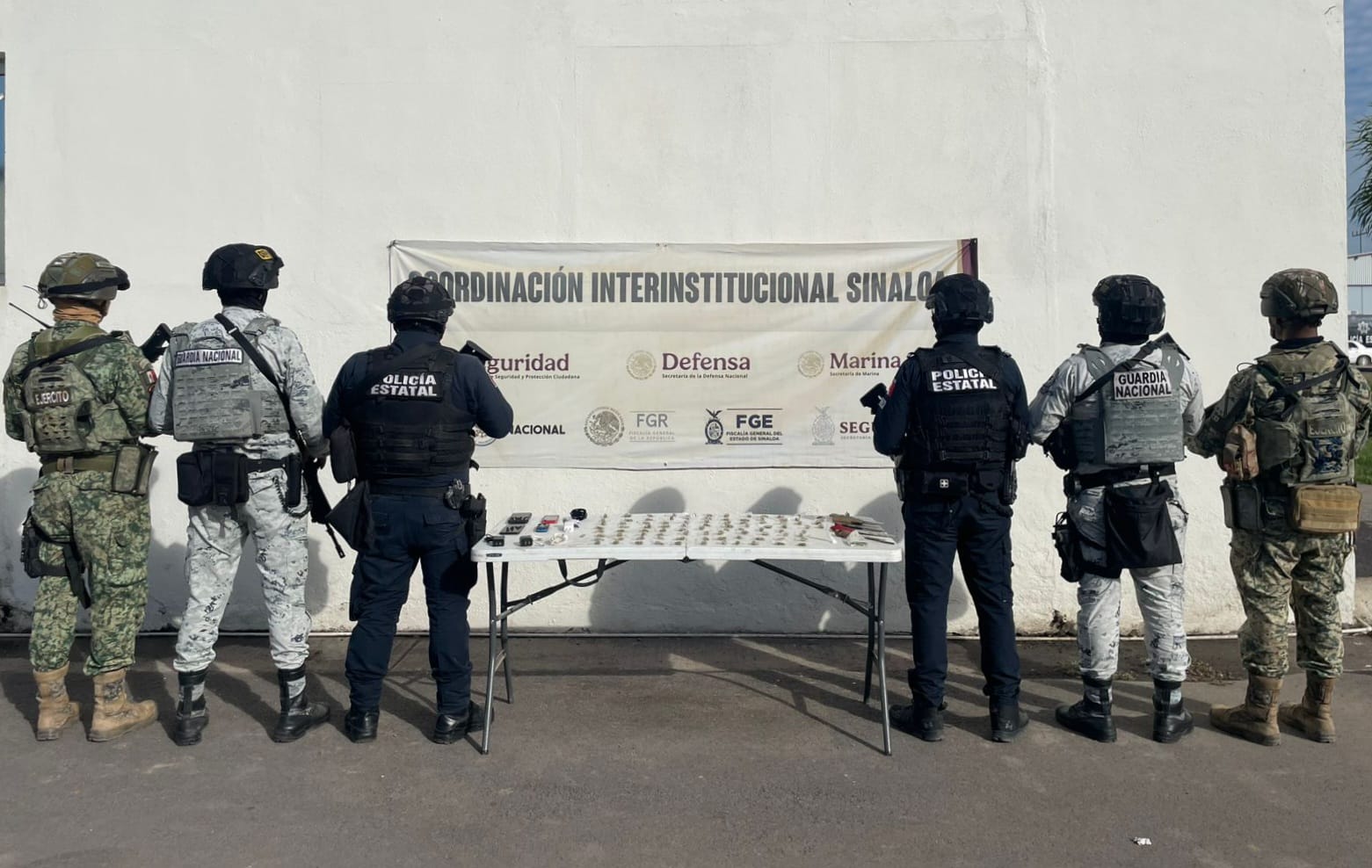 Elementos policiacos junto a una mesa con diferentes objetos y una lona
