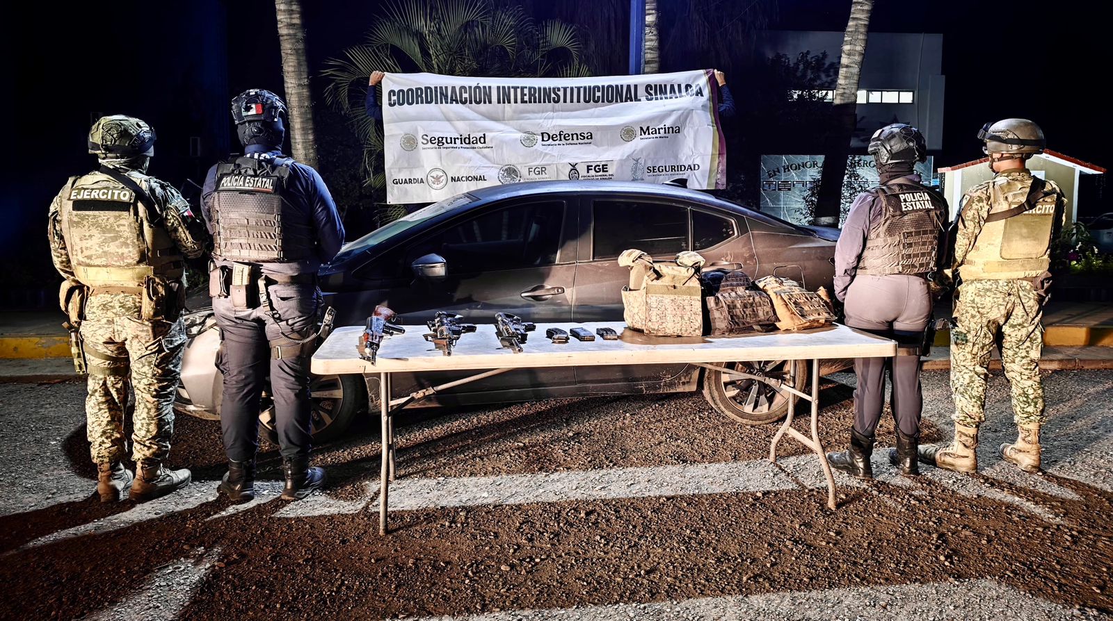 Elementos policiacos junto a una mesa con armas, cargadores y más equipo que fue asegurado, al fondo un vehículo y una lona