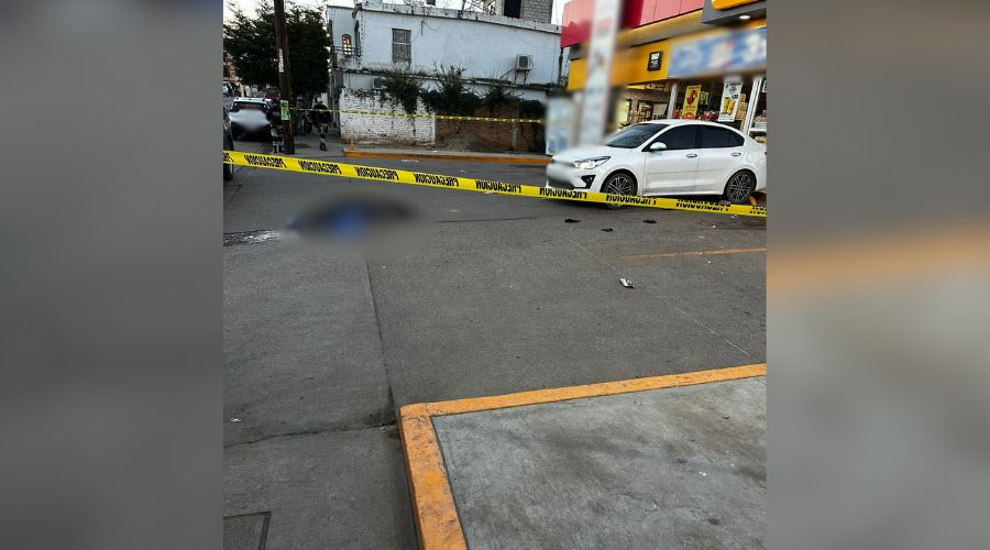 Cuerpo de una persona en el suelo de un estacionamiento de una tienda de autoservicio, se observa un carro blanco y una cinta amarilla de precaución
