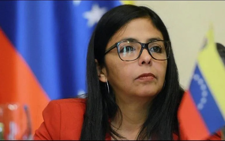 Mujer con lentes viendo hacia el frente con un fondo de una bandera de Venezuela
