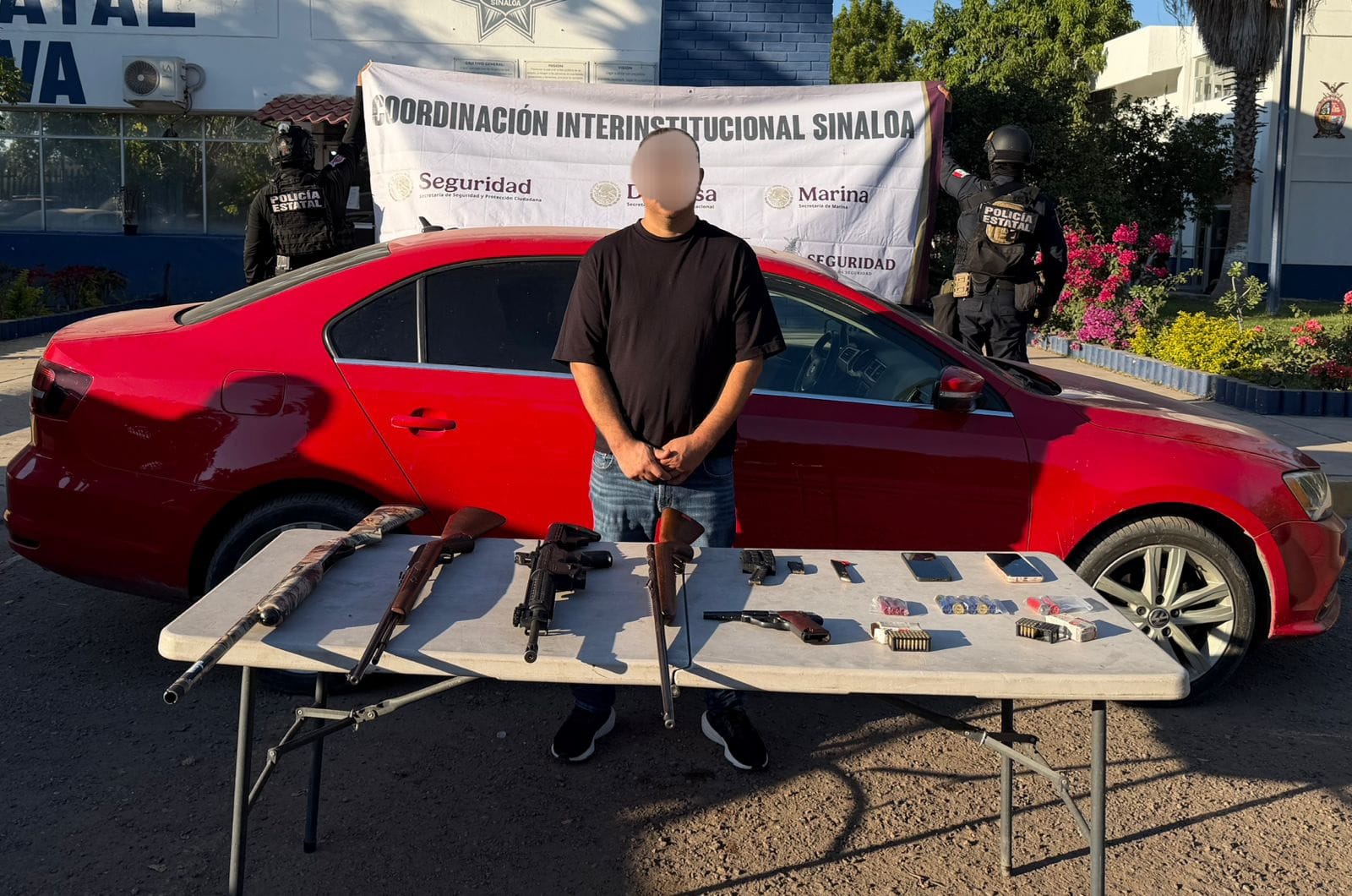 Persona con el rostro cubierto, adelante hay una mesa con armas que fueron aseguradas y al fondo un carro rojo