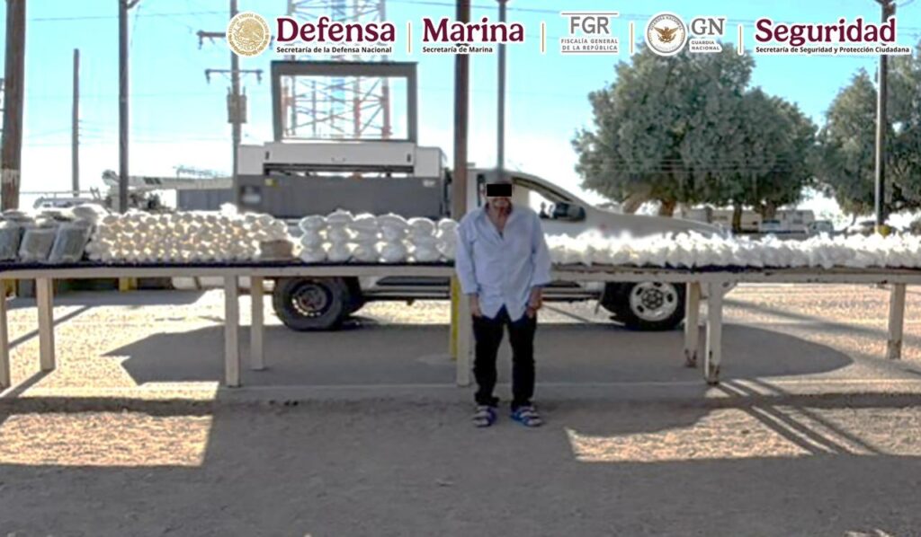 Persona de pie adelante de una mesa con bolsas con un contenido blanco (metanfetamina) y una camioneta asegurada