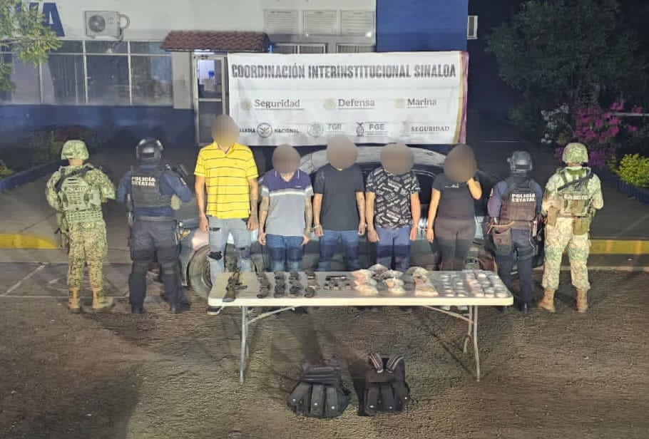 elementos policiacos junto a 5 personas, una mesa con armas y objetos asegurados, al fondo una lona y un automovil también asegurados