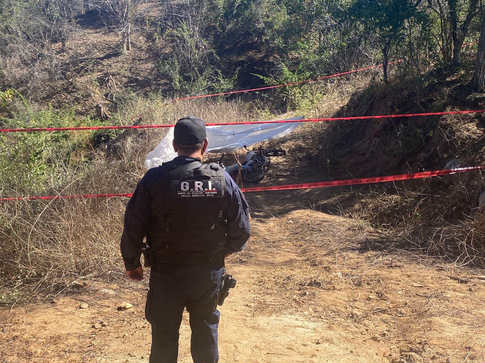 Personal policiaco de espalda frente a una cinta amarilla de precaución, una motocicleta en el suelo y una tela color blanco entre matorrales en un camino de tierra