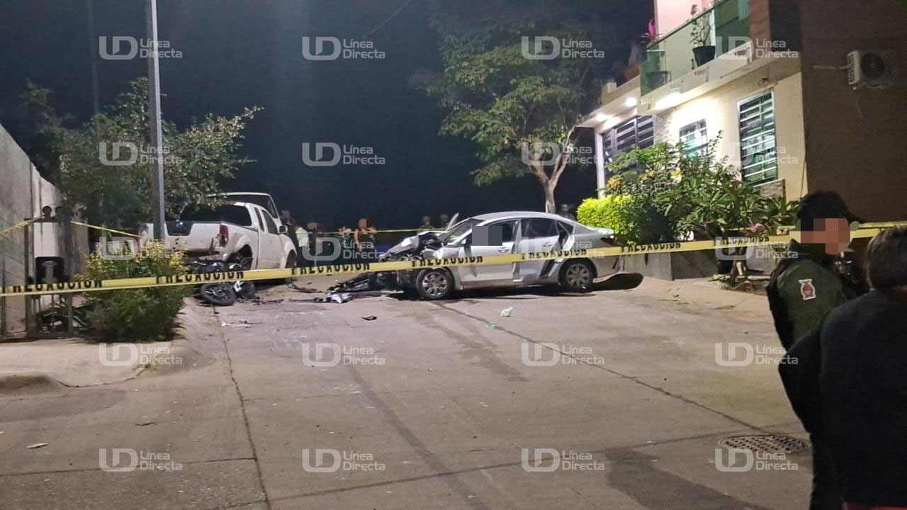 Carro color gris chocado de la parte de enfrente entre una cinta amarilla de precaución en una calle de un vecindario con personas y elementos policiacos alrededor