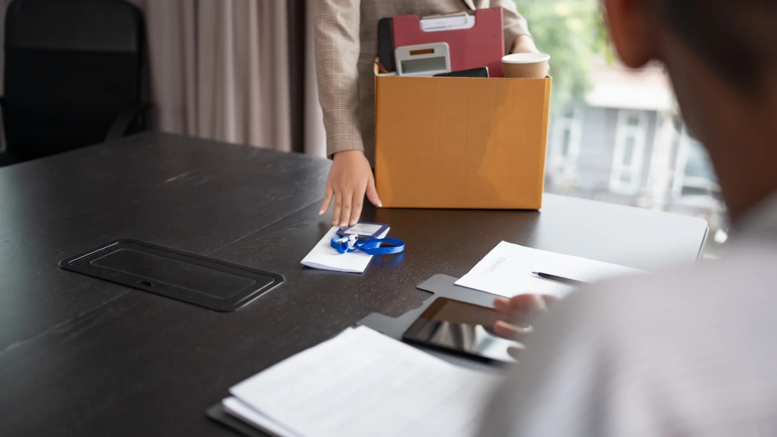 Persona detrás de un escritorio sentada y otra en frente, de pie y con una caja con artículos personales