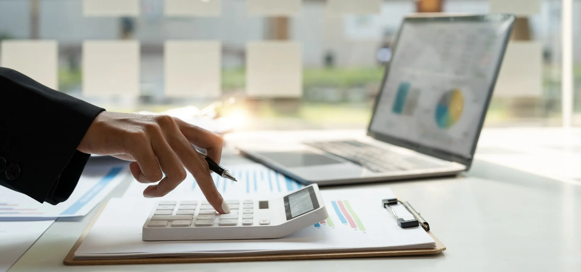 Mano presionando una calculadora y al fondo una laptop con estadísticas