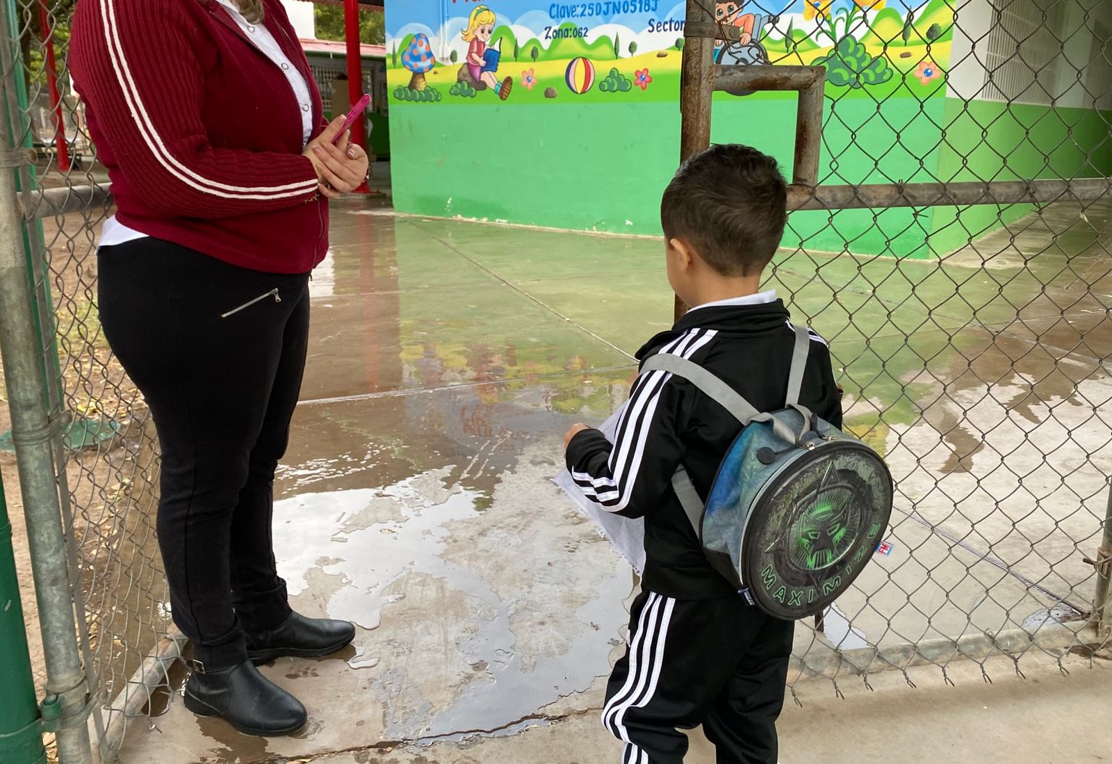 Niño con mochila entrando a una escuela en donde está una persona parada y se observa el suelo mojado por la lluvia