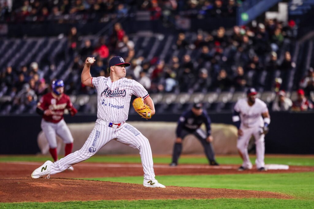 Jugador de beisbol lanzando una pelota en un estadio, al fondo se observan más jugadores