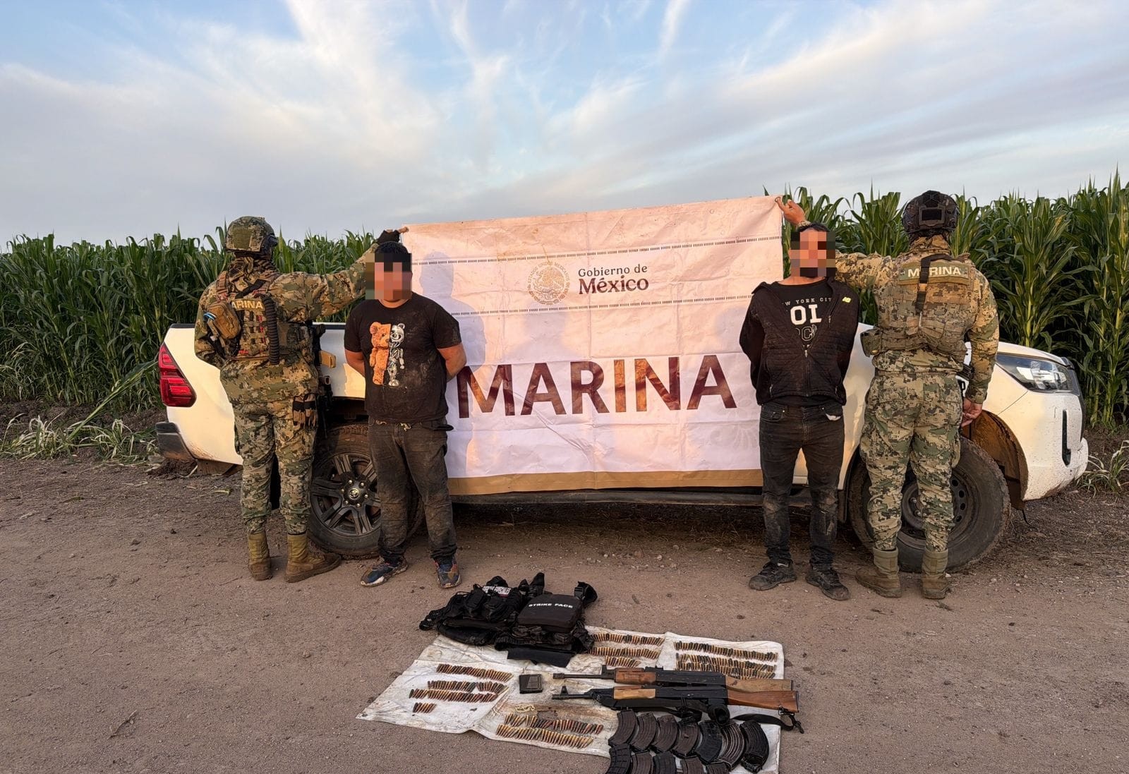 Elementos de la Marina sosteniendo una lona con la leyenda MARINA junto a dos personas detenidas, en el suelo hay otra lona con armas y objetos que fueron asegurados y al fondo se observa un carro blanco