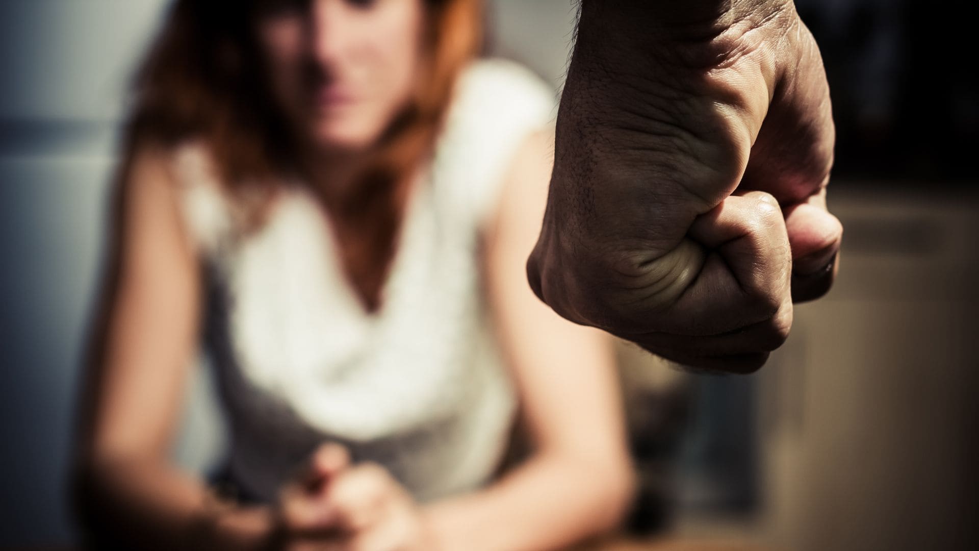 Imagen temática de violencia contra las mujeres con una mujer sentada con una postura de encogimiento y al frente se observa el puño de un hombre