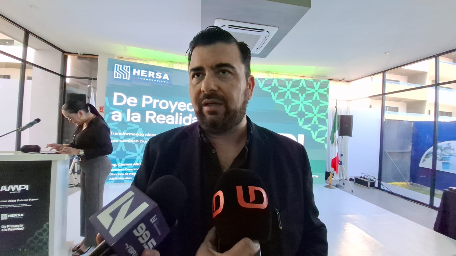 Luciano Carrasco Altamirano vestido de negro hablando a micrófonos fondo azul