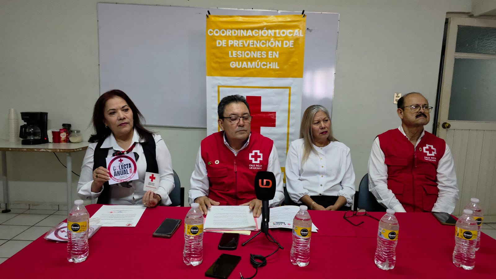 personal de cruz roja sentados detrás de una mesa con lona y pizarrón detrás