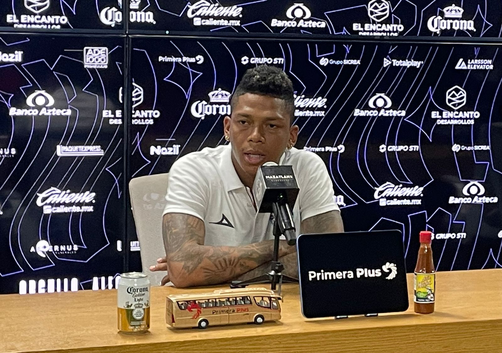 billy arce sentado hablando a un micrófono con lona con nombres de patrocinadores de fondo