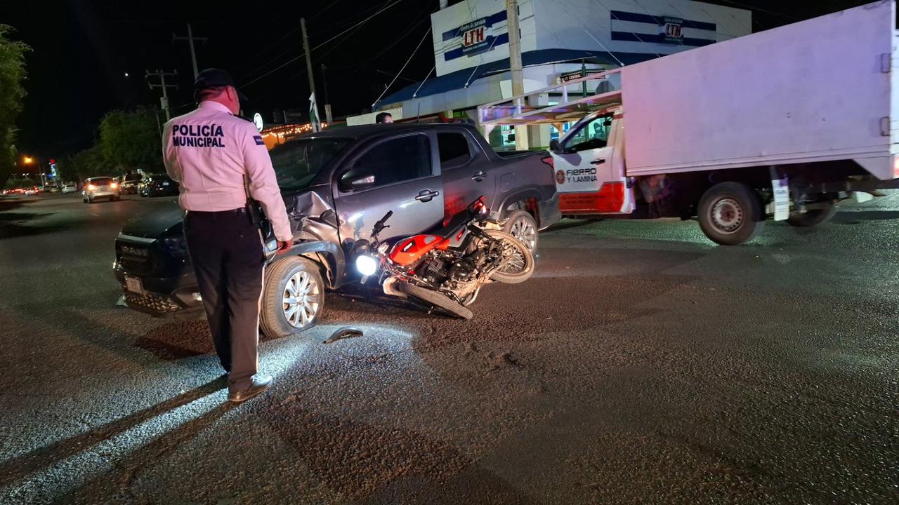 Unidad de transporte gris en medio de la calle con una motocicleta debajo de ella después de un choque y un oficial de transito revisando el accidente