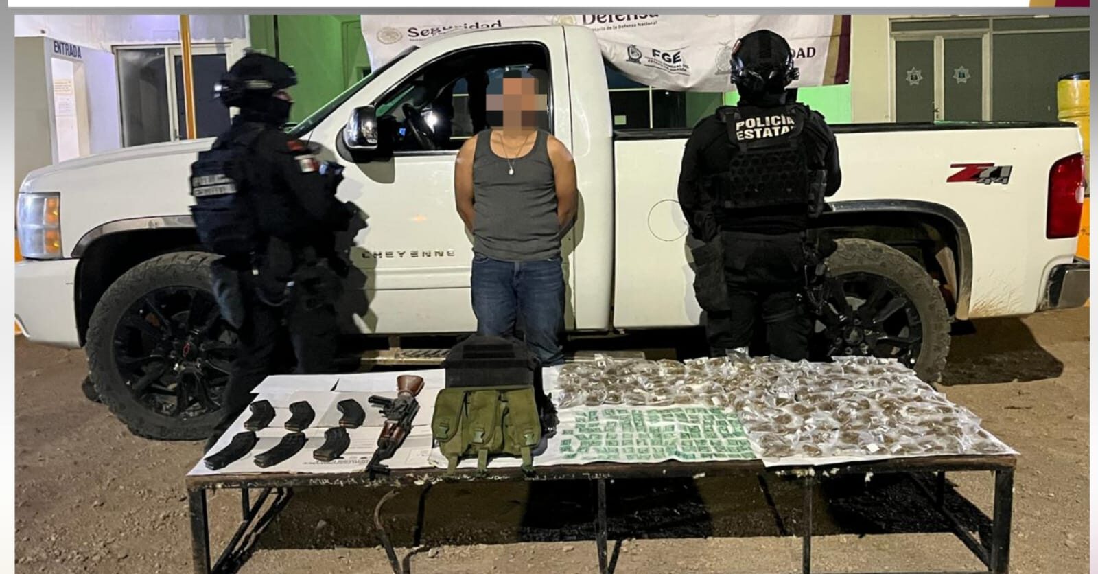 Dos elementos de seguridad junto a una persona detenida y una camioneta con una mesa y armas aseguradas