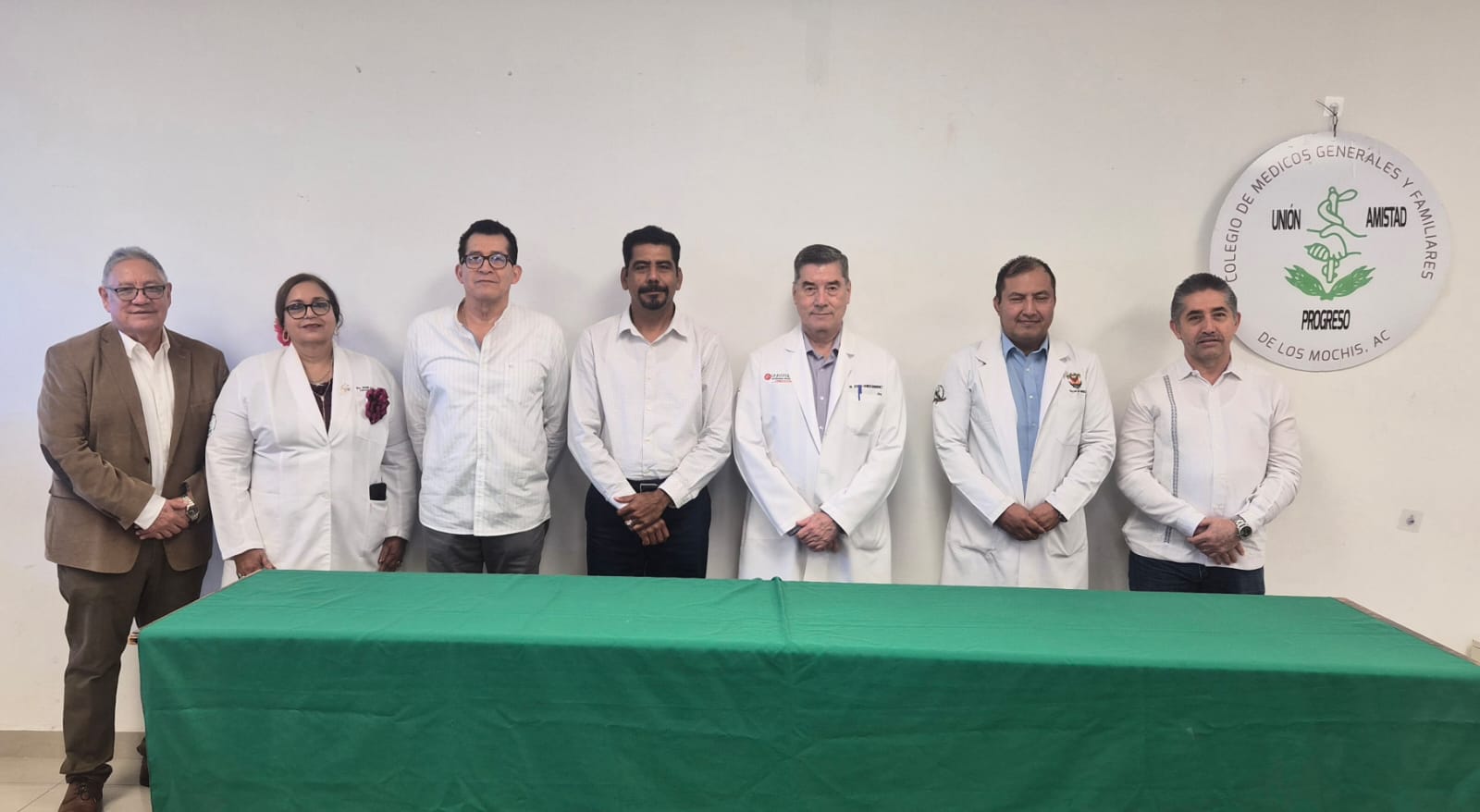 personas de CMGyFLM de pie detrás de mesa con mantel verde y pared blanca de fondo