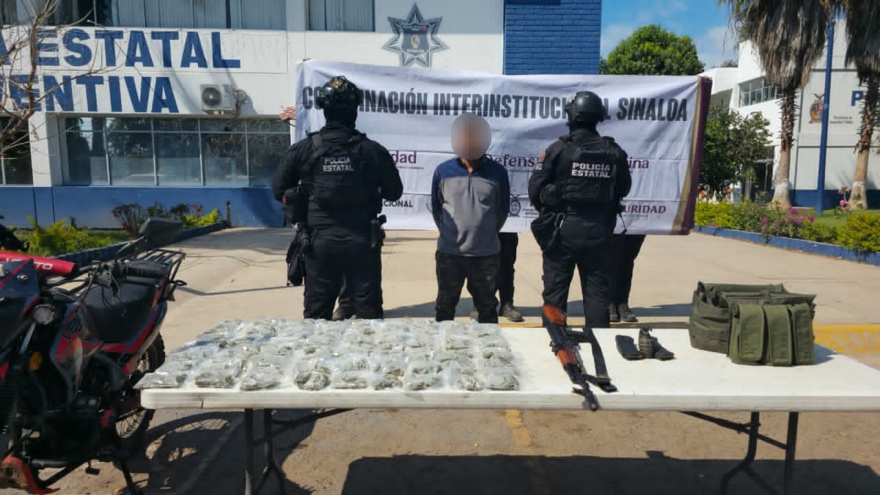 Un hombre con la cara difuminada junto a dos elementos de la policía y objetos asegurados sobre una mesa