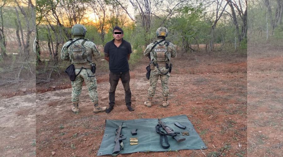 Elementos del Ejécito Mexicano y en el medio una persona detenida, en el suelo una manta y sobre ella artefactos y armas