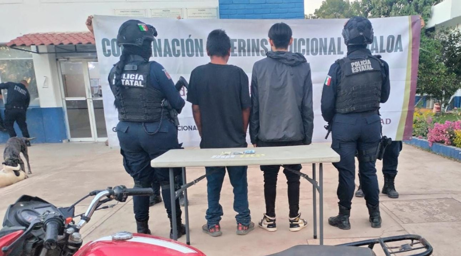 Elementos de seguridad y detenidos de espaldas, una mesa con cosas confiscadas y una motocicleta roja