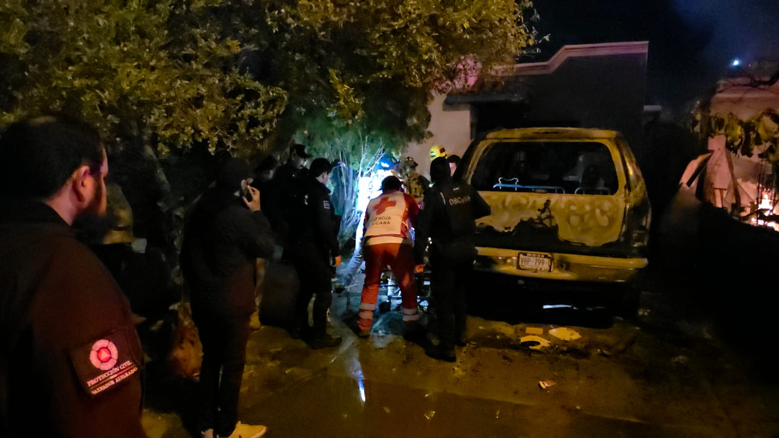 Personas alrededor de una vivienda que está quemada y un auto dañado por el incidente, elemento de la cruz roja auxiliando a alguien