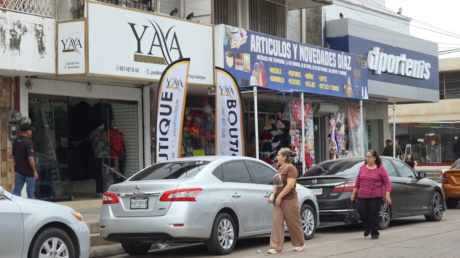 Personas pasando por unos carros y negocios al fondo