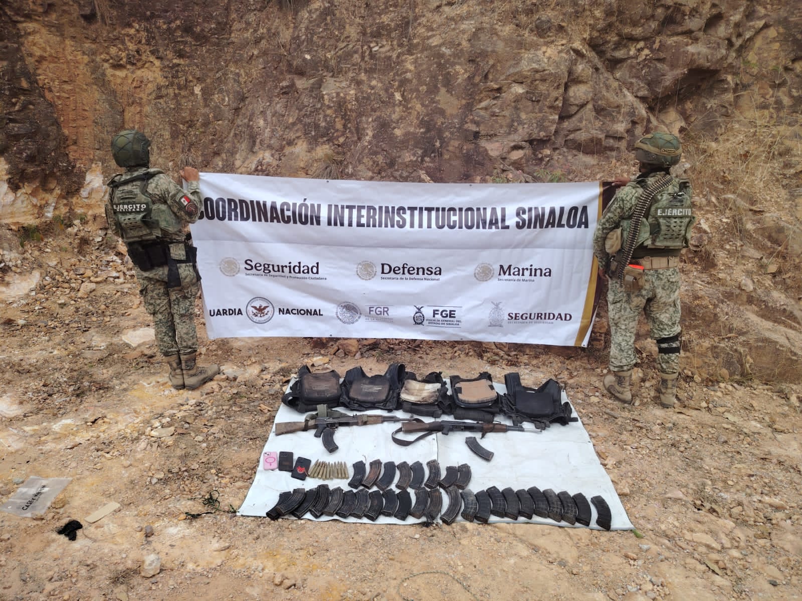 Elementos del ejercito de espaldas sosteniendo una pancarta y en el suelo armas aseguradas en operativo