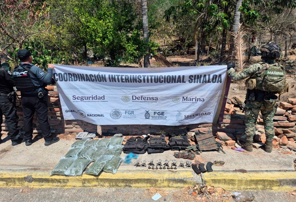 Elementos de seguridad de espaldas sosteniendo una pancarta y debajo de ellos artefactos confiscados