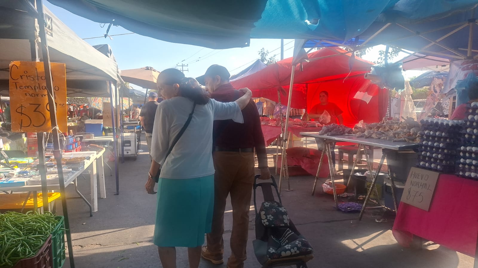 Dos adultos mayores caminando por el centro de un tianguis abrazados