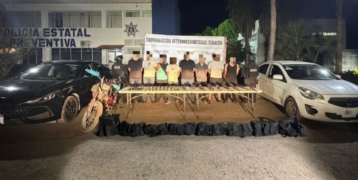 Elementos policiacos junto a personas con las manos en la espalda detenidos y en una mesa objetos y armas aseguradas y dos carros y una motocicleta
