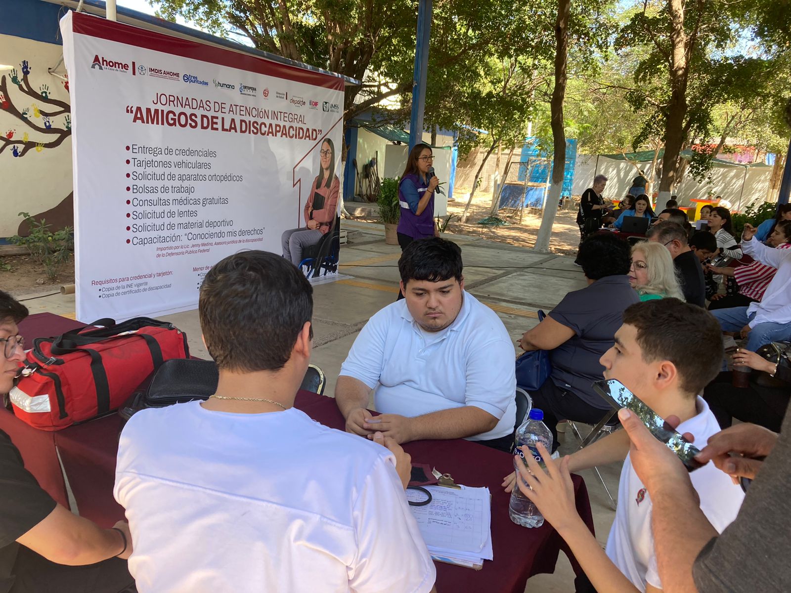 Personas sentadas en un evento, al fondo un templete con la descripción del evento