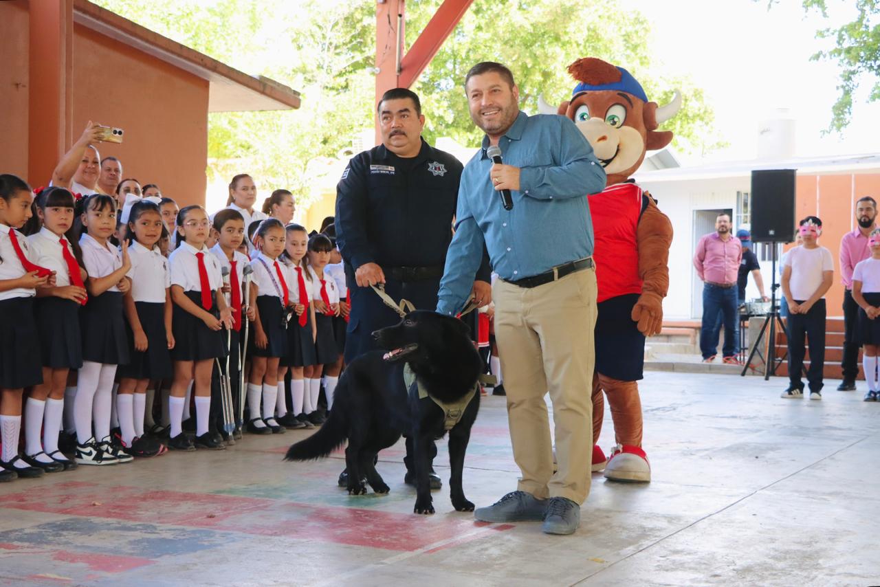 Alcalde de Badiraguato con un micrófono junto a un oficial de policía que sostiene a un perro de vigilancia, todo dentro de una escuela primaria