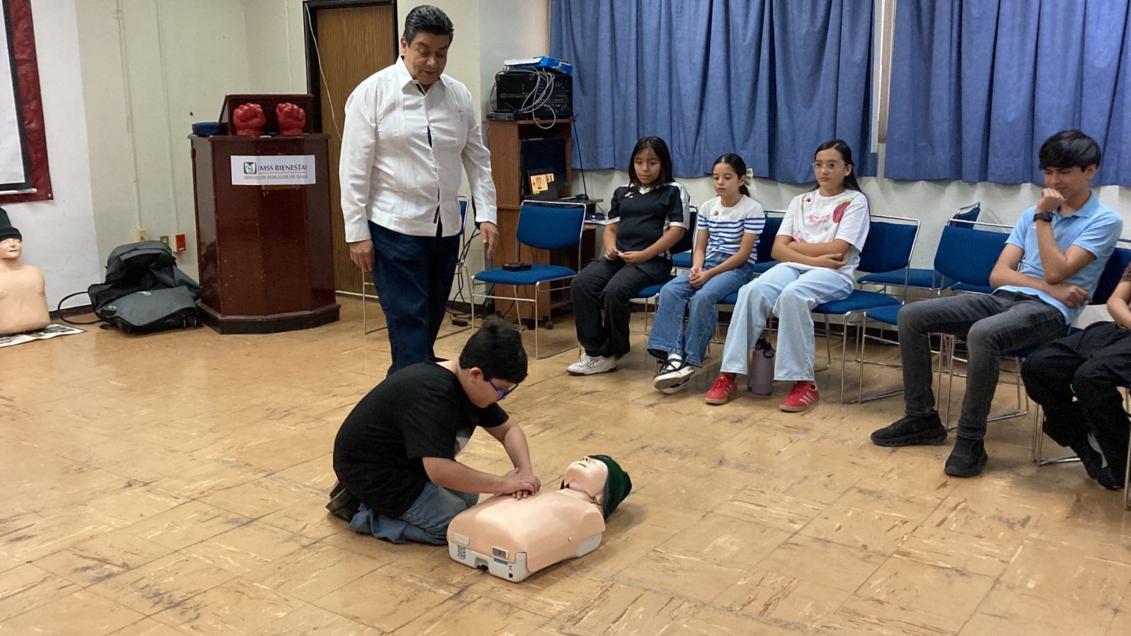 Un niño practica maniobras de reanimación cardiopulmonar en un maniquí mientras personal médico supervisa y otros menores observan durante un taller de RCP en el Hospital General de Los Mochis.