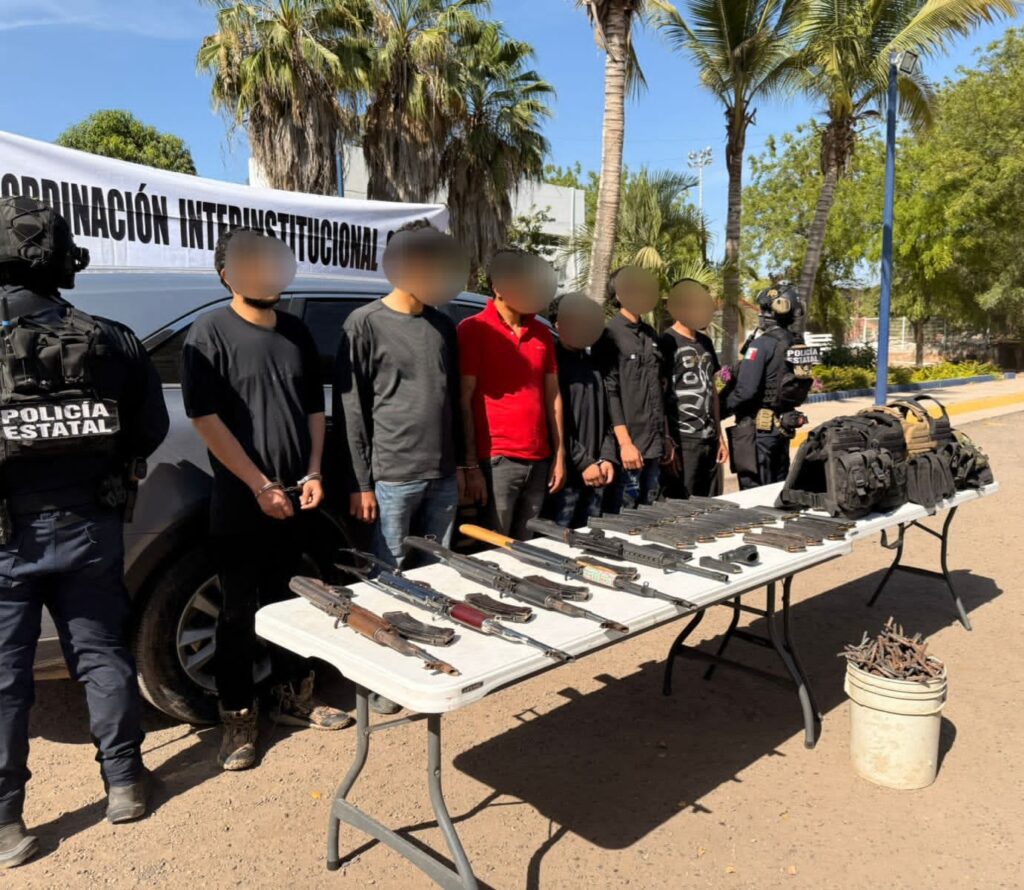 Persona con la cara difuminada y una mesa con armas y una lona de seguridad detrás de las personas