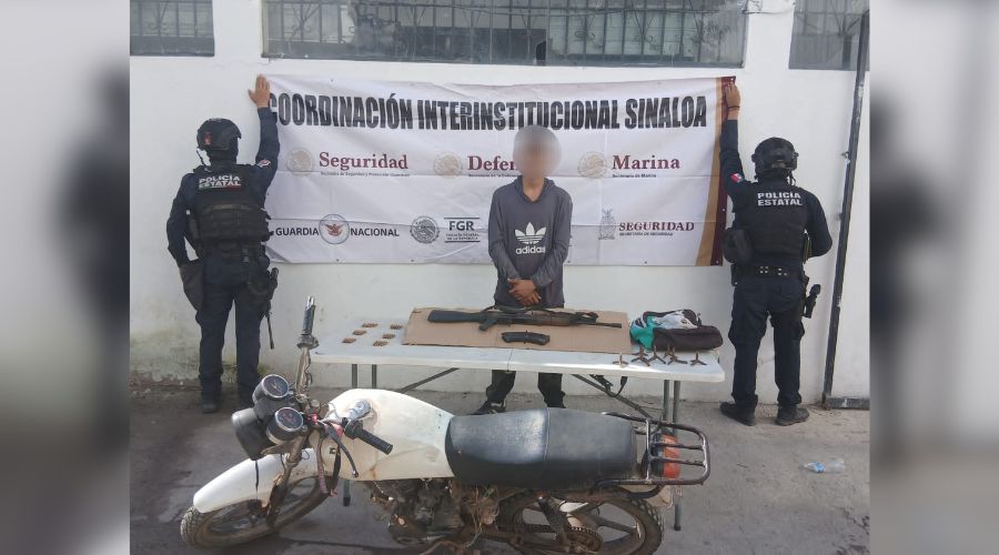 Elementos policiacos de espalda y en el medio una persona detenida y una mesa con objetos y armas y al frente una motocicleta