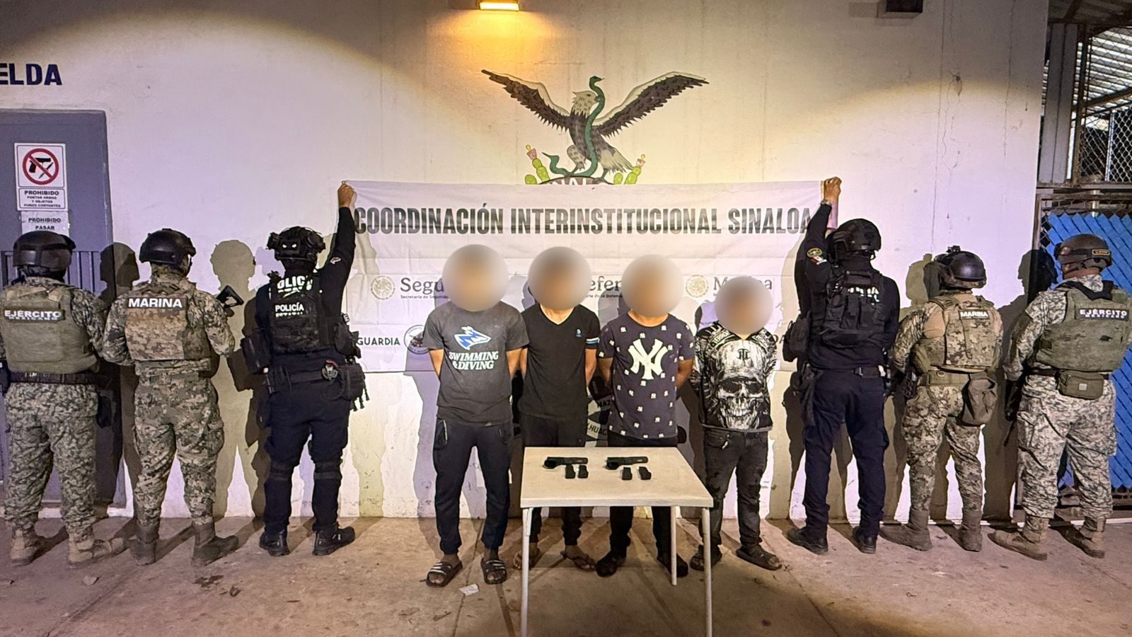 Cuatro personas con el rostro cubierto y elementos policiacos de espaldas en las orillas sosteniendo una lona con logos de corporaciones policiacas