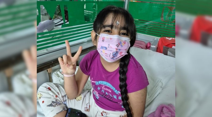 Niña Alexia con cubrebocas en camilla de hospital saludando con su mano derecha