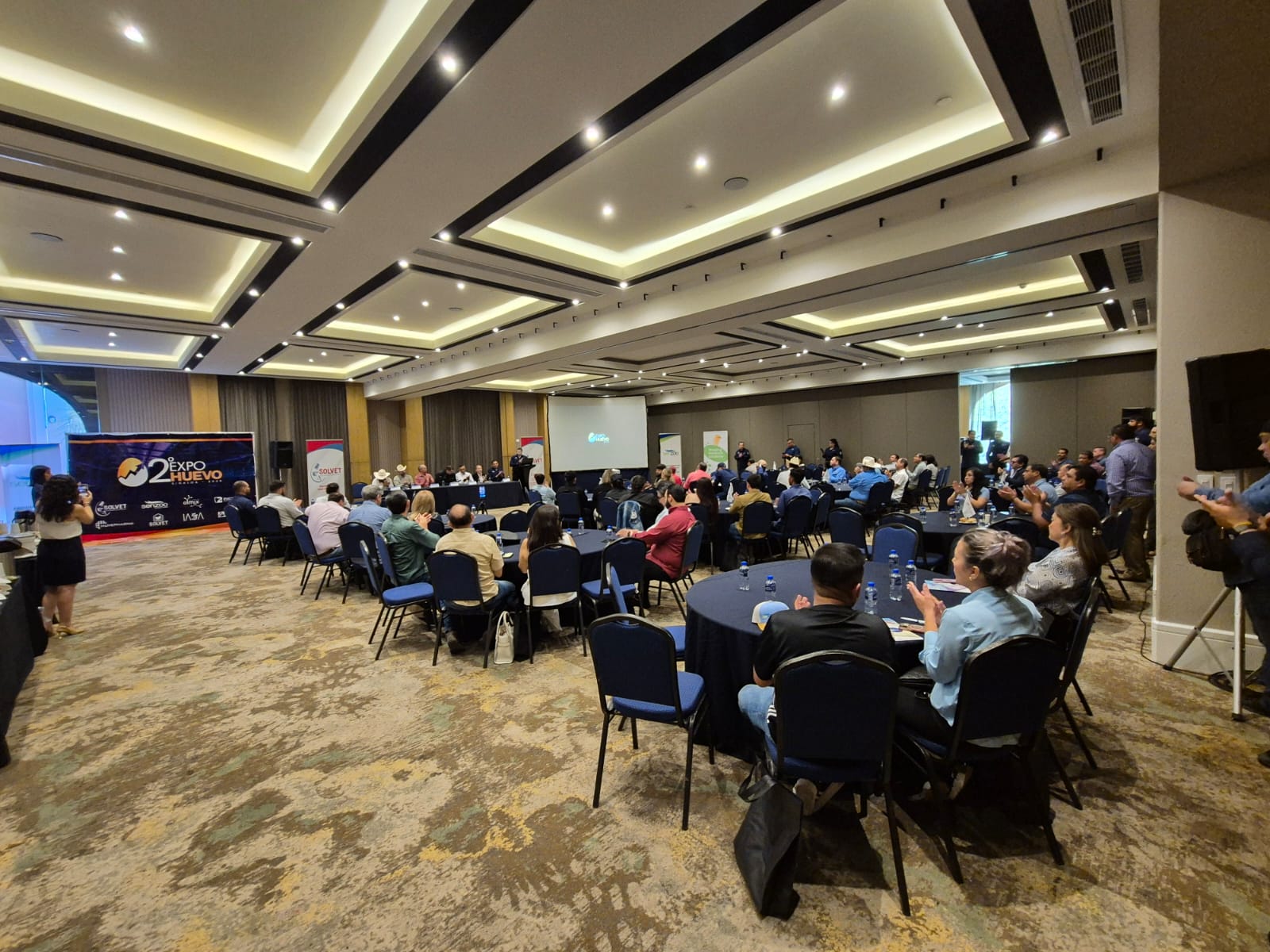 Personas dentro de un salón de eventos y sentados en mesas presenciando una conferencia
