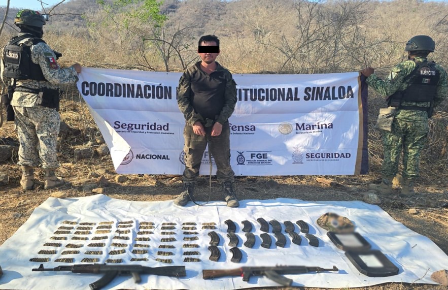 Elementos de seguridad de espaldas sosteniendo una pancarta y en medio una persona detenida con los ojos tapados y debajo de ellos una manta con artefactos confiscados