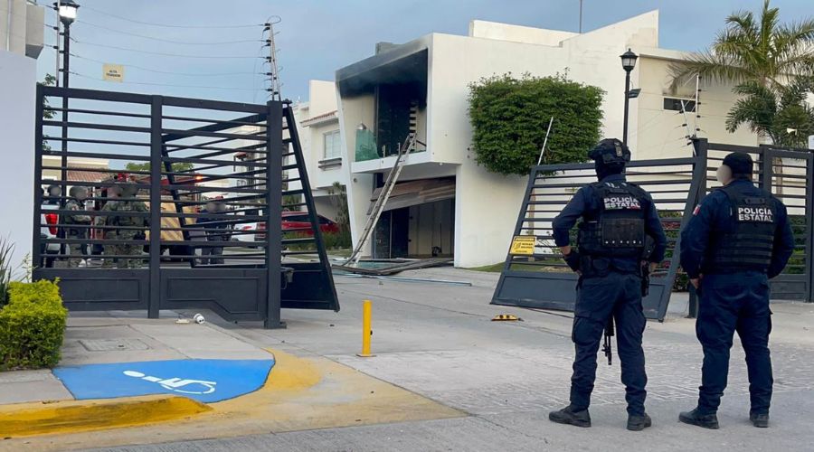 Elementos policiacos desplegados en una zona residencial junto a un portón destruido y una casa con manchas negras tras incendio